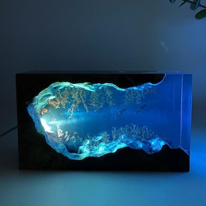 May include: A rectangular resin art piece with an illuminated underwater scene, featuring coral and two fish. The artwork is set within a dark wooden frame. The internal light source creates a blue glow.