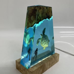 May include: A clear resin sculpture of a sea turtle and two divers, set against a blue background. The sculpture is mounted on a wooden base.