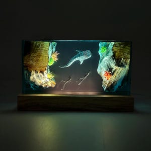 Puede incluir: Una escultura de resina de un tiburón ballena nadando en un océano azul con dos buceadores. La escultura está iluminada desde atrás, creando un efecto brillante. La escultura está sobre una base de madera.
