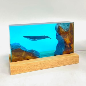 Puede incluir: Una pieza de arte rectangular de resina que representa una ballena nadando en un océano azul. La obra de arte está encapsulada en resina transparente con detalles marrones y azules, colocada sobre una base de madera natural. La ballena es gris oscuro.