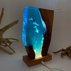 May include: A blue resin lamp with a wooden base features a 3D image of whales swimming in the ocean. The lamp is lit up with a warm white light.