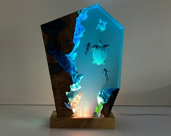 May include: A resin sculpture of a blue ocean scene with a large sea turtle, two smaller sea turtles, and two scuba divers. The sculpture is lit from behind, creating a glowing effect. The sculpture is mounted on a wooden base.