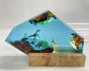 May include: A unique resin art piece featuring two dinosaur figurines set within a light blue, transparent enclosure. The piece includes wood and faux greenery, mounted on a wooden base. The overall design is a decorative, artistic display.