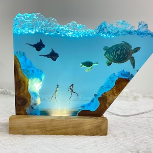 May include: A unique resin art piece depicting an underwater scene with sea turtles, stingrays, and divers. The artwork is illuminated with blue LED lights, set on a wooden base, creating a captivating ocean-themed display. The piece is a decorative accent.