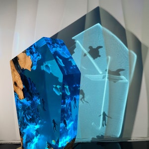 May include: A blue resin sculpture with a wooden base, featuring a scene of sea turtles swimming in a blue ocean. The sculpture is illuminated from within, creating a glowing effect.