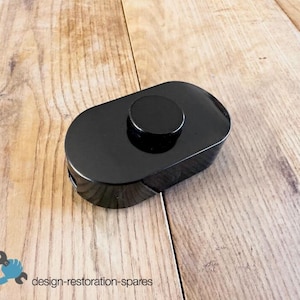 Op de afbeelding: Een zwarte, ovale voetschakelaar met een ronde knop aan de bovenkant. De glanzende schakelaar ligt op een houten oppervlak. De tekst "design-restoration-spares" is linksonder zichtbaar.
