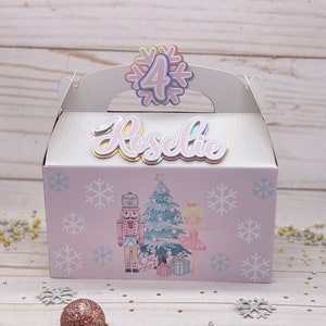May include: A pink and white favor box with a snowflake design and a ballerina and nutcracker illustration. The box has a handle and says "Roselie" and "4" on the front.