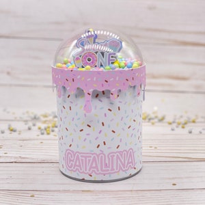 May include: A pink and white cylinder shaped container with a clear plastic dome on top. The container has a sprinkle pattern and the words "Sweet One" and "Catalina" are printed on it. The dome contains colorful sprinkles and a small plastic ice cream cone with a pink and white donut on top.