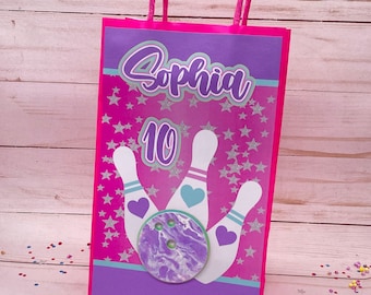 May include: Pink and purple goody bag with a bowling theme. The bag has a pink background with silver stars and a purple border. It says "Sophia 10" and has a bowling ball and pins.