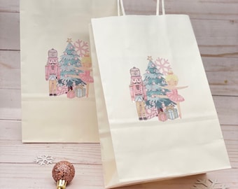 May include: Two white paper gift bags with handles. Each bag features a colorful illustration of a nutcracker, a ballerina, and a Christmas tree. The bags are on a light pink wooden surface with gold and silver glitter snowflakes.