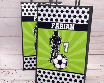Op de afbeelding: Zwarte en groene cadeauzakken met een voetbalthema met een zwart-wit voetbalpatroon. De tas heeft een groene achtergrond met een silhouet van een voetballer en de naam "FABIAN" en het nummer "7" erop gedrukt.