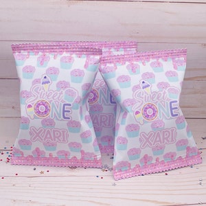 May include: Two pink and white treat bags with a cupcake pattern. The bags have a pink and white frosting design with sprinkles. The bags say "Sweet One" and "Xari" in purple text.