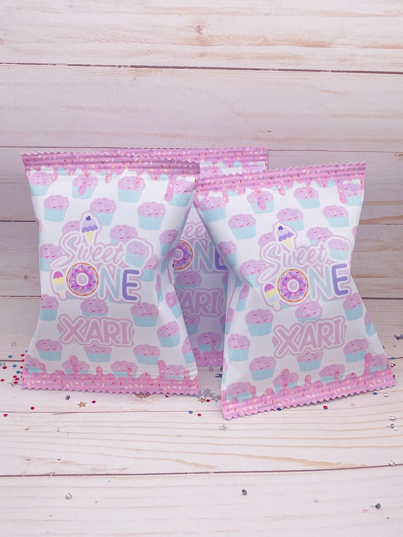 May include: Two pink and white treat bags with a cupcake pattern. The bags have a pink and white frosting design with sprinkles. The bags say "Sweet One" and "Xari" in purple text.