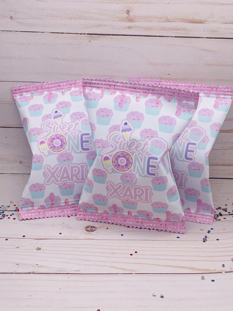 May include: Three pink and white treat bags with a cupcake and donut pattern. The bags have the text "Sweet One" and "Xari" printed on them.