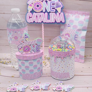 May include: Sweet One party decorations with a pink, purple, and blue color scheme. The decorations include a cake topper, cupcake toppers, a water bottle label, a favor bag, and a favor box. The decorations feature a donut, ice cream cone, and cupcake design. The text "Sweet One Catalina" is featured on the cake topper and favor box.