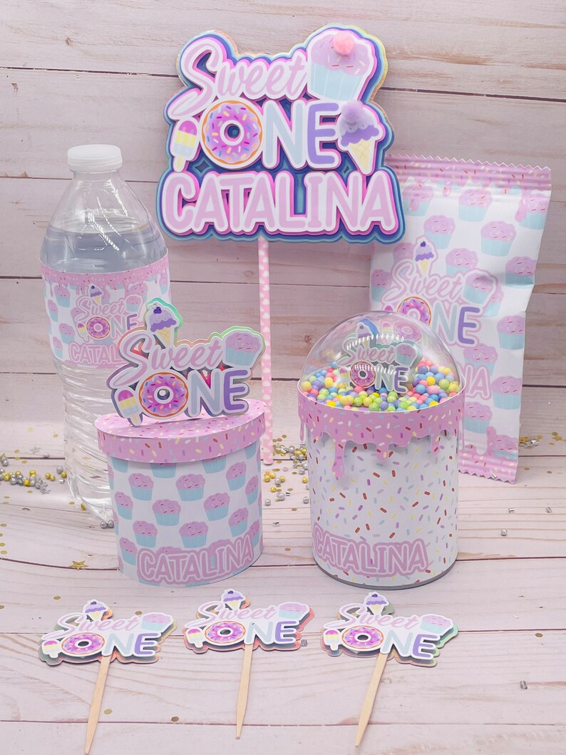 May include: Sweet One party decorations with a pink, purple, and blue color scheme. The decorations include a cake topper, cupcake toppers, a water bottle label, a favor bag, and a favor box. The decorations feature a donut, ice cream cone, and cupcake design. The text "Sweet One Catalina" is featured on the cake topper and favor box.