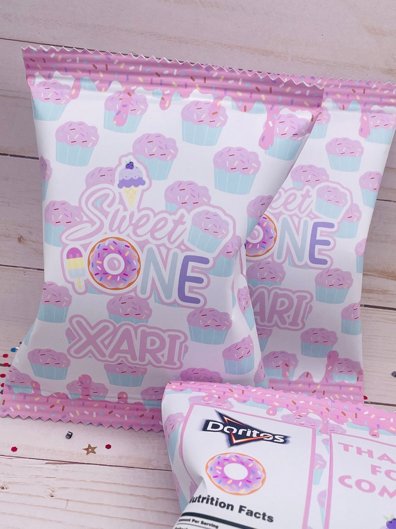 May include: Two pink and white bags with a donut and cupcake pattern. The bags have the text "Sweet One Xari" on them. The bags are filled with Doritos.