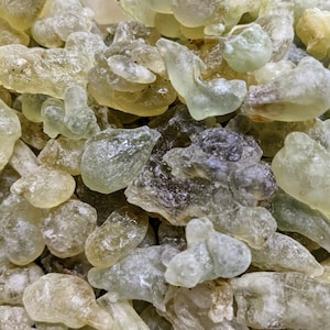 May include: A close-up of a pile of translucent, light green and brown resin pieces. The resin is irregularly shaped and has a slightly rough texture.