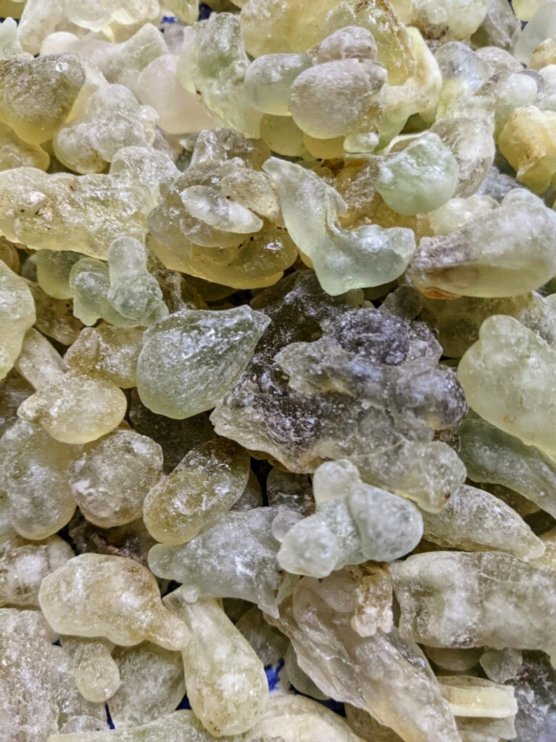 May include: A close-up of a pile of translucent, light green and brown resin pieces. The resin is irregularly shaped and has a slightly rough texture.