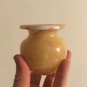Puede incluir: Un pequeño jarrón de alabastro amarillo con un borde blanco. El jarrón está siendo sostenido en la mano de una persona.