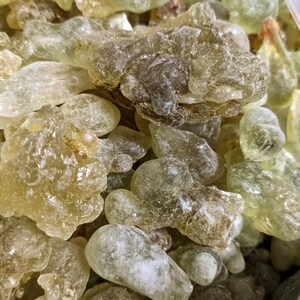 May include: Close-up of a pile of translucent, light green and brown resin pieces. The resin is irregularly shaped and has a slightly rough texture.