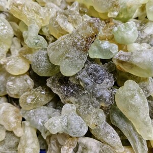 May include: A close-up of a pile of translucent green and brown resin pieces. The resin is irregularly shaped and has a rough, textured surface.