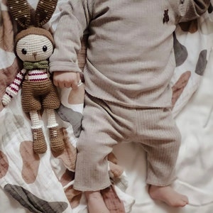 May include: A brown crocheted bunny toy with red and white stripes on its sleeves and a green bow tie lies on a white and brown patterned blanket next to a baby wearing a brown ribbed long-sleeve shirt and pants.