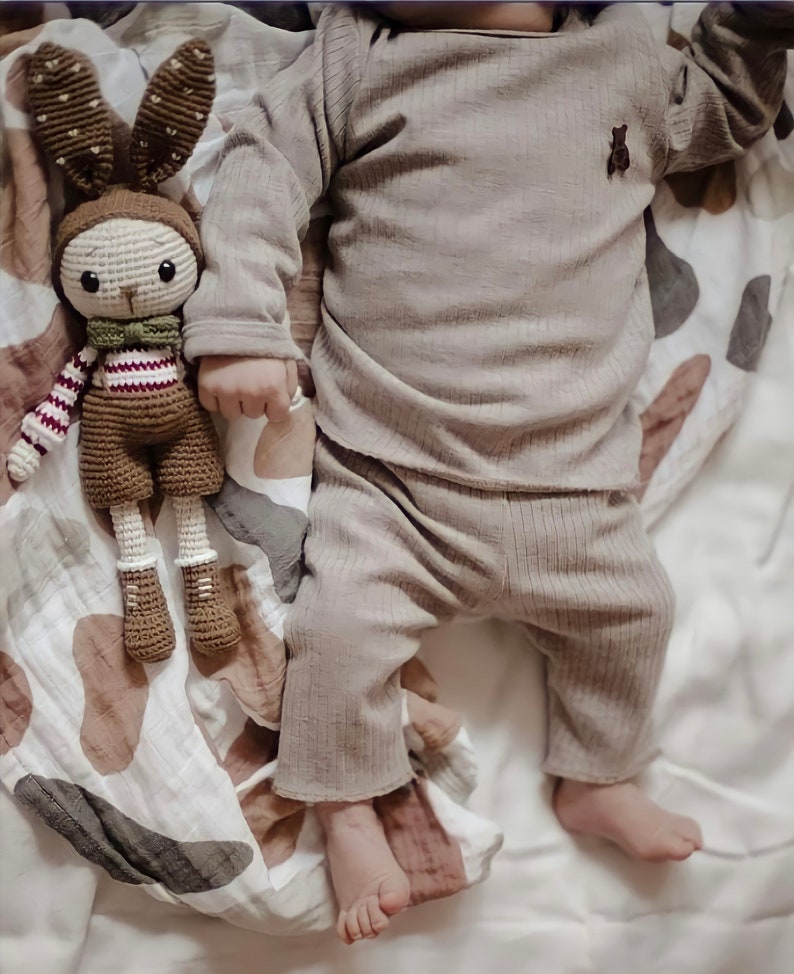 May include: A brown crocheted bunny toy with red and white stripes on its sleeves and a green bow tie lies on a white and brown patterned blanket next to a baby wearing a brown ribbed long-sleeve shirt and pants.