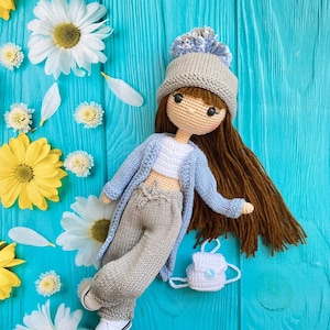 Puede incluir: Una muñeca de crochet con un gorro de punto gris, una chaqueta azul, pantalones grises y zapatillas blancas. La muñeca tiene el pelo castaño y lleva una pequeña mochila blanca de crochet. La muñeca está tumbada sobre una superficie de madera azul con flores blancas.