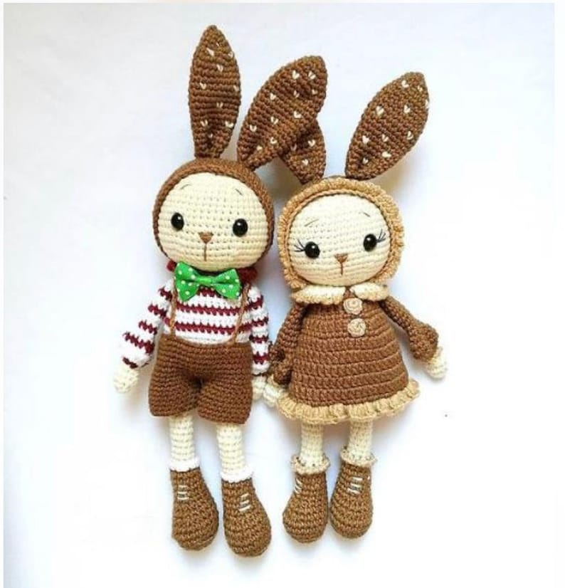May include: Two crocheted bunny dolls, one wearing a brown dress with a white collar and the other wearing a striped shirt and brown pants. Both dolls have brown ears with white polka dots.