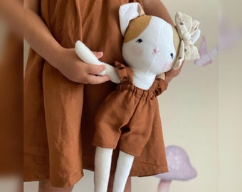 Op de afbeelding: Een witte en bruine kattenpop die een bruine overalljurk draagt met een witte ruche en een bruine strik. De pop wordt vastgehouden door een kind dat een bruine jurk draagt.