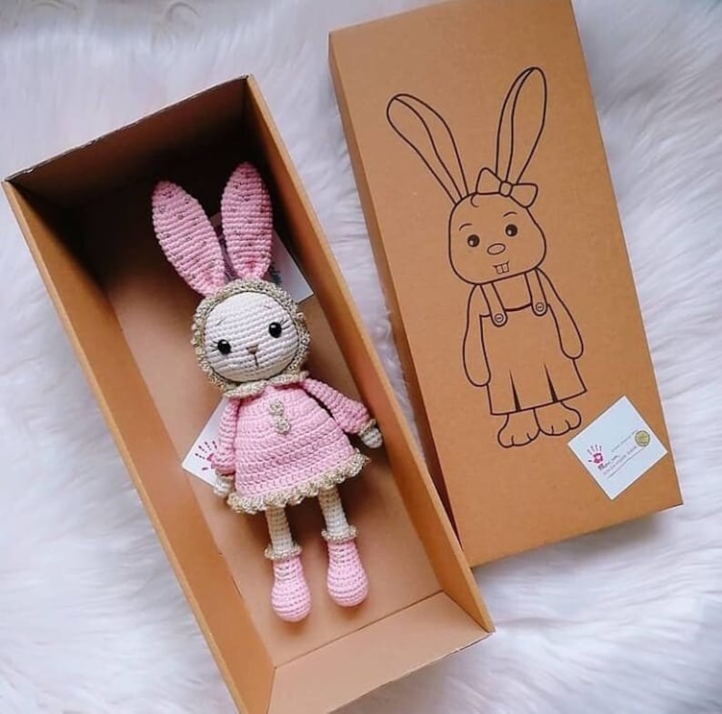May include: A pink crocheted bunny rabbit toy wearing a pink dress and gold trim. The bunny is in a brown cardboard box with a smaller brown box featuring a line drawing of a bunny on the side.