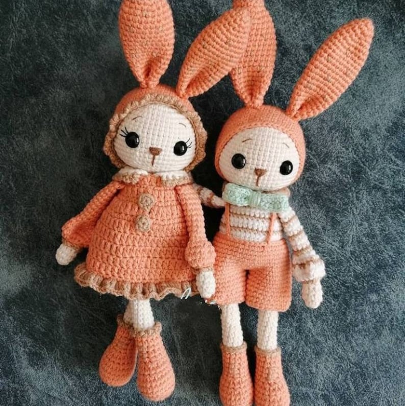 May include: Two crocheted bunny dolls, one wearing a peach dress and the other wearing peach overalls with white stripes. Both dolls have peach ears and white faces with black eyes.