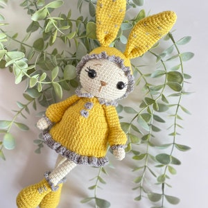 May include: A crocheted yellow bunny rabbit wearing a grey and white dress. The bunny has large black eyes and a small brown nose.