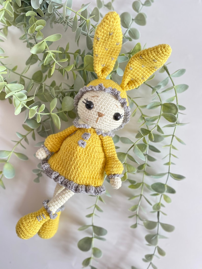May include: A crocheted yellow bunny rabbit wearing a grey and white dress. The bunny has large black eyes and a small brown nose.