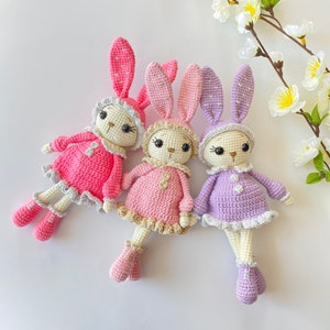 May include: Three crocheted bunny dolls in pink, purple, and light pink with white accents. The bunnies are wearing dresses and have embroidered eyes and noses.