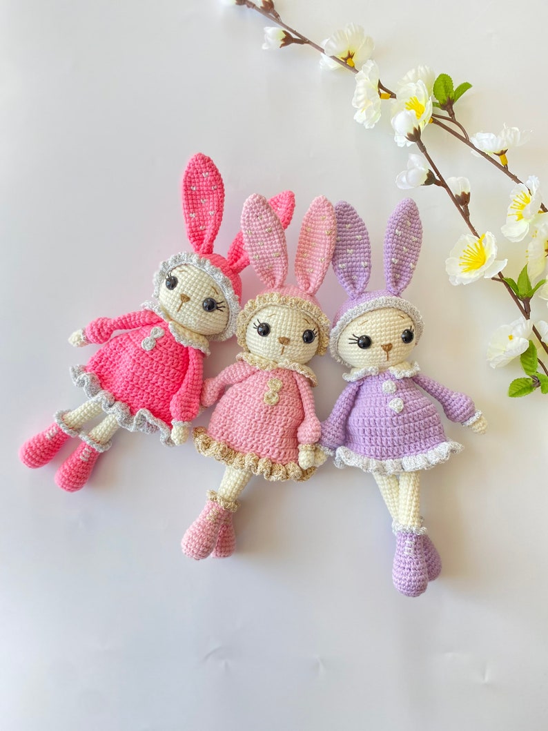 May include: Three crocheted bunny dolls in pink, purple, and light pink with white accents. The bunnies are wearing dresses and have embroidered eyes and noses.