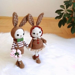 May include: Two crocheted bunny dolls, one brown with a green bow tie and a striped shirt, the other brown with a red hood and a brown dress. The dolls are standing on a white fluffy surface.