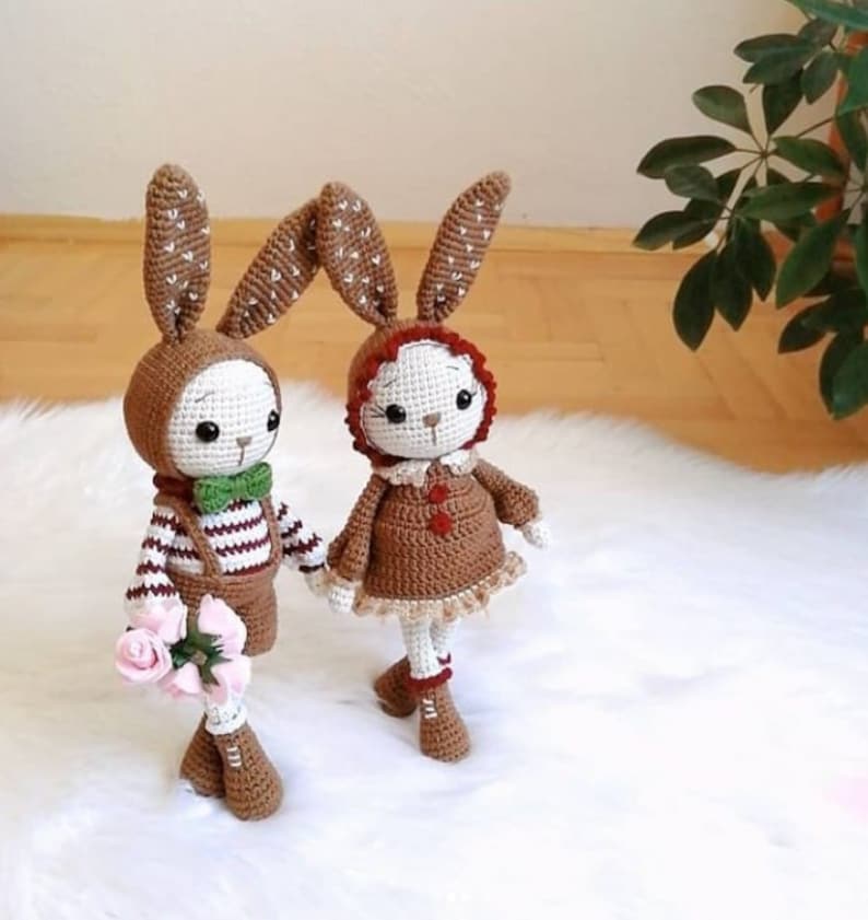 May include: Two crocheted bunny dolls, one brown with a green bow tie and a striped shirt, the other brown with a red hood and a brown dress. The dolls are standing on a white fluffy surface.