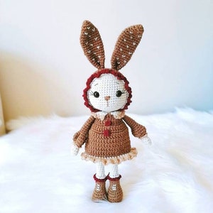 May include: A crocheted bunny doll wearing a brown dress with a red trim and brown boots. The bunny has white fur and brown ears with white polka dots.
