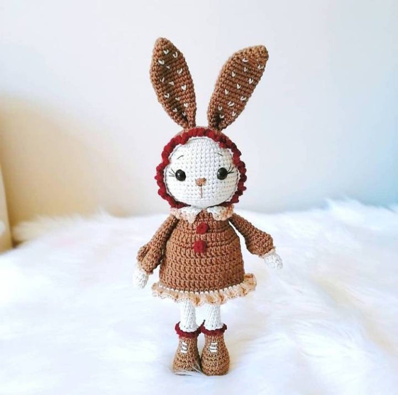May include: A crocheted bunny doll wearing a brown dress with a red trim and brown boots. The bunny has white fur and brown ears with white polka dots.