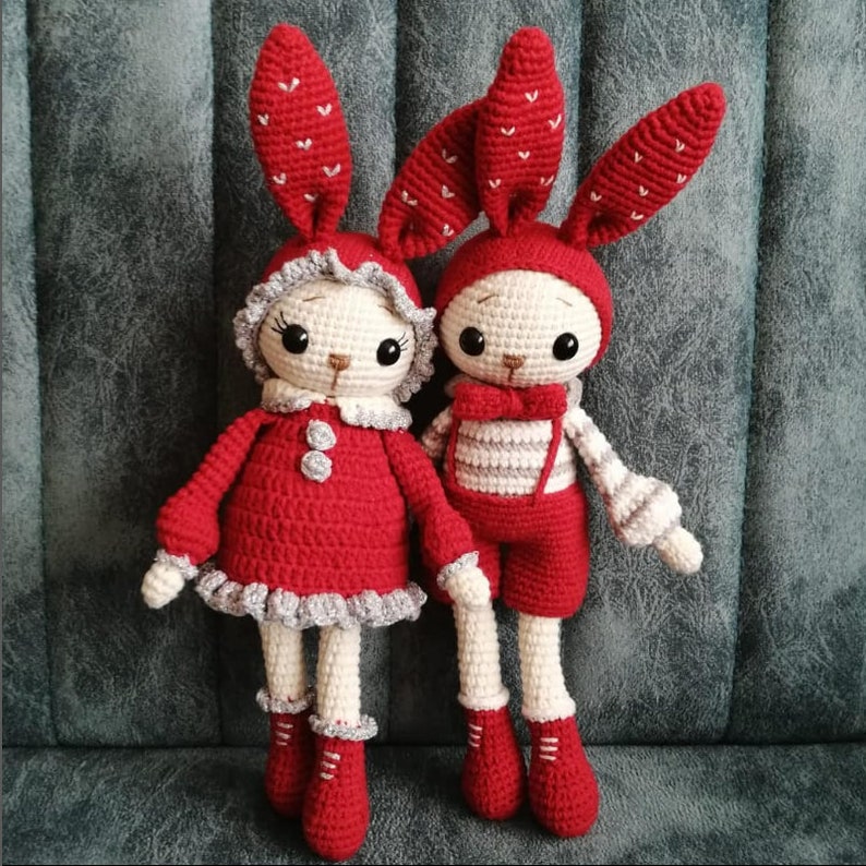 May include: Two crocheted bunny dolls with red and white striped outfits. The bunny on the left is wearing a red dress with white trim and the bunny on the right is wearing a white and red striped shirt and red pants.
