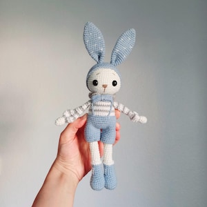 May include: A crocheted blue bunny toy with white and gray striped overalls. The bunny has long white legs and blue ears with white polka dots.