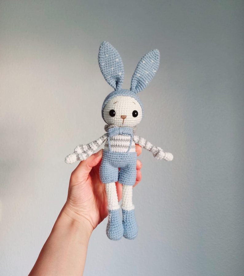 May include: A crocheted blue bunny toy with white and gray striped overalls. The bunny has long white legs and blue ears with white polka dots.
