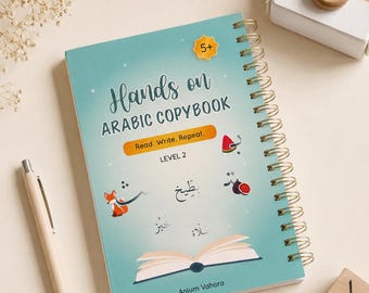 May include: A spiral-bound "Hands on ARABIC COPYBOOK" with a teal cover, featuring Arabic script and illustrations. The book is labeled "Level 2" and includes the text "Read. Write. Repeat." A pen, wooden blocks, and a toy camera are also present.