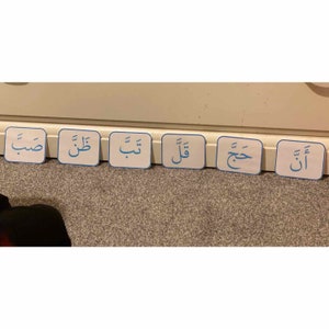 Puede incluir: Seis tarjetas cuadradas blancas con bordes azules y escritura &aacute;rabe en azul. Las tarjetas est&aacute;n dispuestas en fila sobre una superficie marr&oacute;n claro. La escritura &aacute;rabe en las tarjetas dice: "أ", "ت", "ح", "و", "ل", "ب".