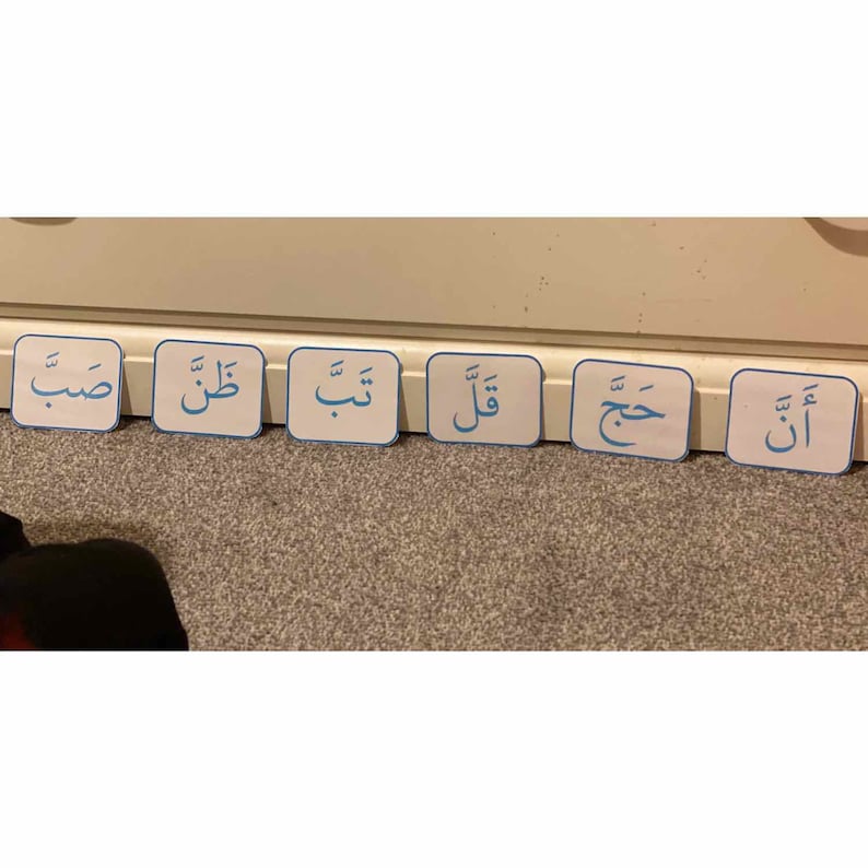 Puede incluir: Seis tarjetas cuadradas blancas con bordes azules y escritura &aacute;rabe en azul. Las tarjetas est&aacute;n dispuestas en fila sobre una superficie marr&oacute;n claro. La escritura &aacute;rabe en las tarjetas dice: "أ", "ت", "ح", "و", "ل", "ب".