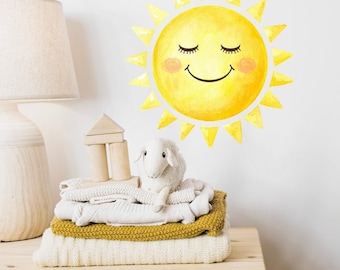 May include: A cheerful yellow sun wall decal with a smiling face and closed eyes. The sun is surrounded by yellow rays. A stack of folded blankets in cream and mustard yellow, a wooden toy house, and a white stuffed sheep are on a wooden surface.