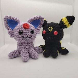 Puede incluir: Dos peluches de crochet, uno morado con orejas azules y nariz roja, el otro negro con detalles amarillos y ojos rojos.