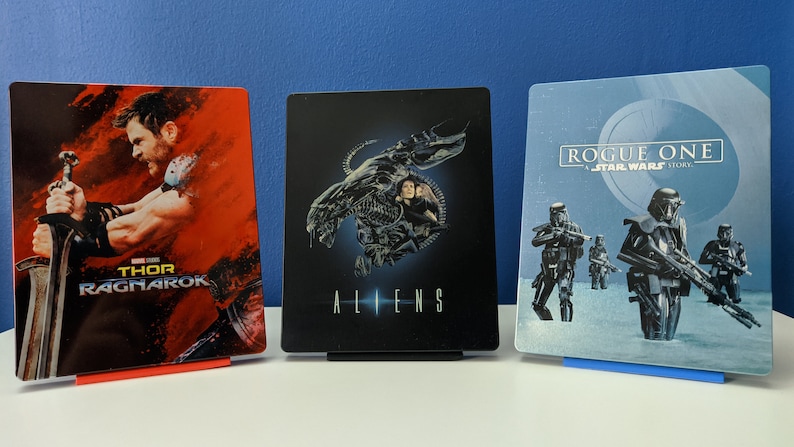 May include: Three steel cases for Blu-ray discs featuring movie posters. The first case is red with a poster for "Thor: Ragnarok" featuring Thor holding a sword. The second case is black with a poster for "Aliens" featuring an alien creature. The third case is blue with a poster for "Rogue One: A Star Wars Story" featuring Stormtroopers.